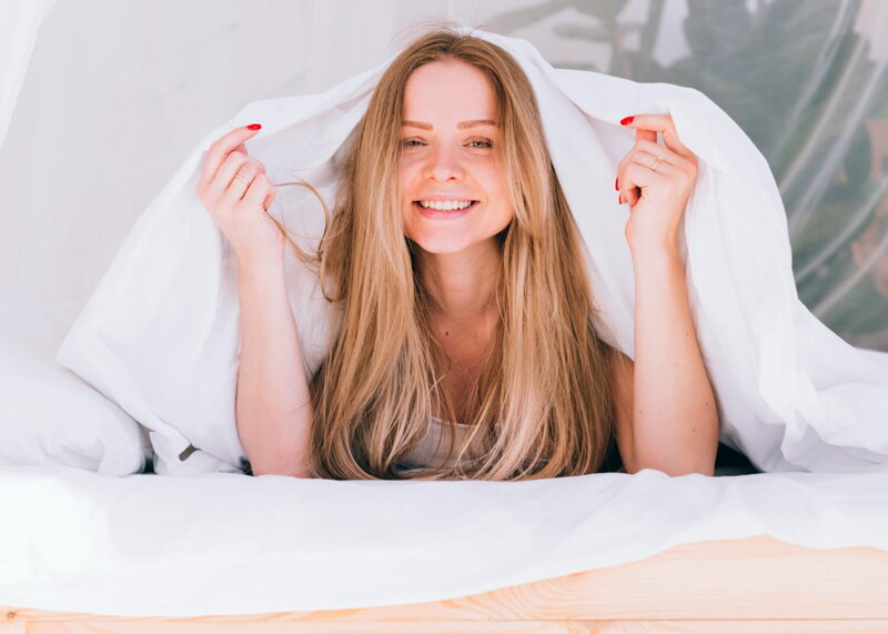 mladá usmiata žena je zakrytá pod žiarivo bielymi perinami